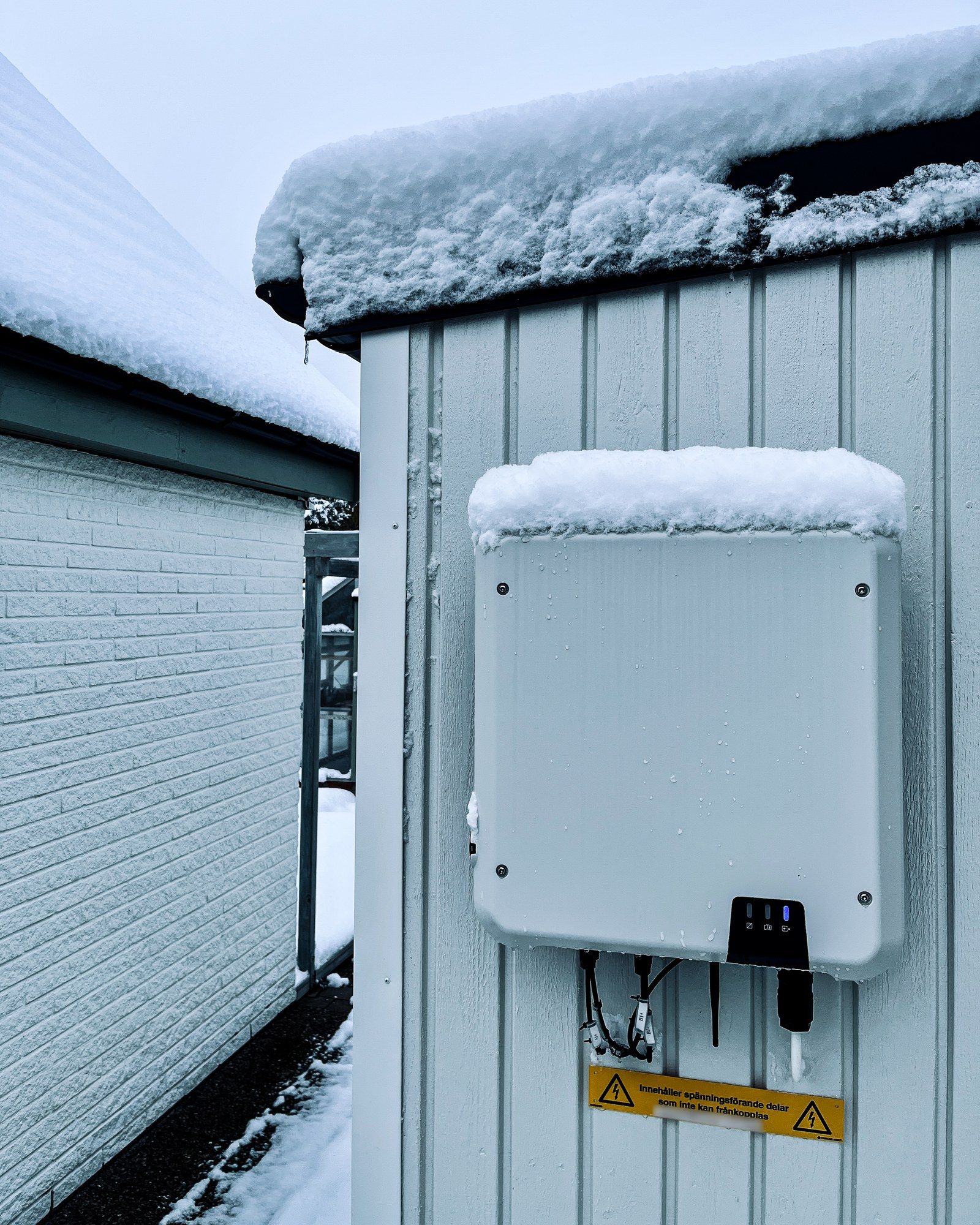Vit växelriktare monterad på en ljus träfasad, med snö på taket och ovanpå apparaten